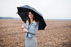 Porträt eines brünetten lockigen Mädchens in Jeansjacke mit schwarzem Regenschirm auf dem Feld. foto