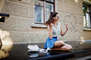 Lockige, stilvolle Mädchenbekleidung auf Blue Jeans-Rock, Bluse und Sonnenbrille. Porträt auf den Straßen der Stadt. foto