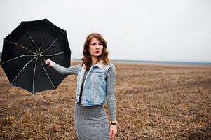 Porträt eines brünetten lockigen Mädchens in Jeansjacke mit schwarzem Regenschirm auf dem Feld. foto