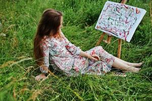 Porträt einer wunderschönen, glücklichen jungen Frau in schönem Kleid, die auf dem Gras sitzt und mit Wasserfarben auf Papier malt. foto