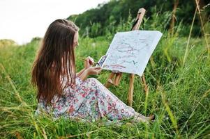 Porträt einer wunderschönen, glücklichen jungen Frau in schönem Kleid, die auf dem Gras sitzt und mit Wasserfarben auf Papier malt. foto