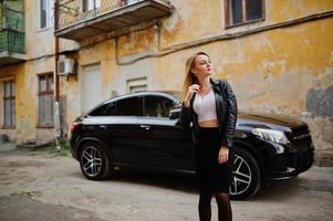 elegantes blondes mädchen trägt eine schwarze lederjacke, die auf den straßen des luxusautos im stadthintergrund posiert. foto