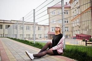 Blondes Mädchen mit Brille und rosa Mantel, schwarze Tunika, die auf der Straße am Zaun sitzt. foto