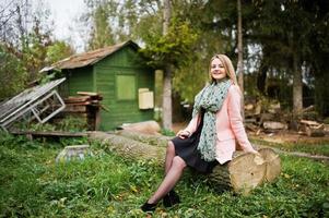 junges blondes mädchen im rosa mantel posierte im herbstpark und saß auf einem gefällten baum gegen das haus des försters. foto