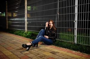 Nachtporträt von Mädchen Model Wear auf Jeans und Lederjacke gegen Eisenzaun. foto