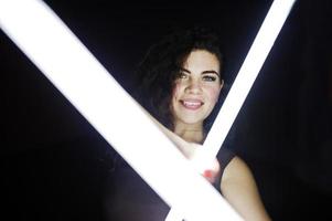 lockiges brünettes plus-size-model mit zwei langen led-lampen im schwarzen raum. foto