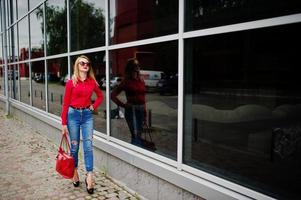 Porträt einer fabelhaften jungen Frau in roter Bluse und Jeans, die mit ihrer Handtasche und Sonnenbrille vor dem Einkaufszentrum auf Glashintergrund posiert. foto