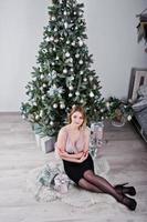 blondes mädchen im kleid posierte in der nähe des neujahrsbaums mit weihnachtsgeschenkdekor im weißen raum. foto