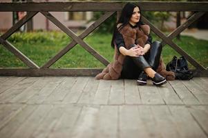 Modefoto im Freien einer wunderschönen sinnlichen Frau mit dunklem Haar in eleganter Kleidung und luxuriösem Pelzmantel, die an Holzgeländern sitzt. foto