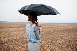 Porträt eines brünetten lockigen Mädchens in Jeansjacke mit schwarzem Regenschirm auf dem Feld. foto