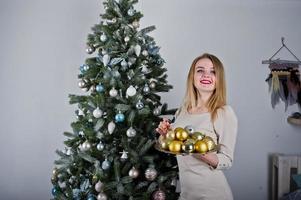 Süßes blondes Mädchen auf beigem Kleid gegen Neujahrsbaum mit goldenen Neujahrsbällen im Studio. schöne Ferien. foto