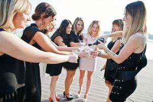 Gruppe von 8 Mädchen tragen auf Schwarz und 2 Bräute beim Junggesellinnenabschied gegen sonnigen Strand, der Champagner trinkt. foto