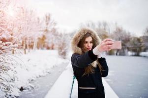 schönes brünettes Mädchen in warmer Winterkleidung. Modell auf Winterjacke gegen zugefrorenen See im Park, der Selfie am Telefon macht. foto