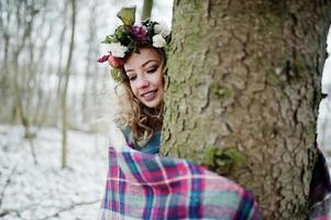 lockiges süßes blondes Mädchen mit Kranz im karierten Plaid am verschneiten Wald am Wintertag. foto