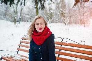 Blondes Mädchen in rotem Schal und Mantel, das am Wintertag auf der Bank sitzt. foto