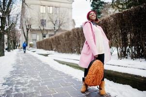 afroamerikanisches Mädchen mit rotem Hut und rosafarbenem Mantel mit Rucksack auf der Straße der Stadt am Wintertag. foto