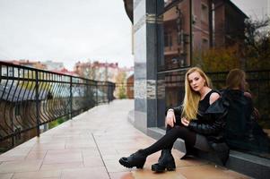 blondes modisches mädchen im langen schwarzen ledermantel, der gegen großes fenster des gebäudes gestellt wird. foto