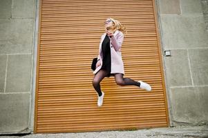 blondes mädchen mit brille und rosa mantel mit handtasche springen gegen fensterläden. foto