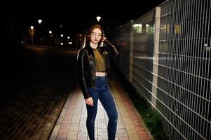 Nachtporträt von Mädchen Model Wear auf Jeans und Lederjacke gegen Eisenzaun. foto