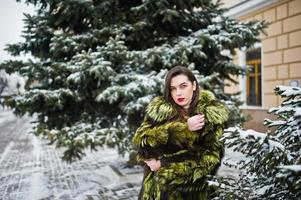 Brunettemädchen im grünen Pelzmantel am Wintertag gegen schneebedeckte Kiefer. foto