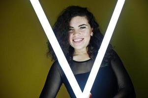 lockiges brünettes plus-size-modell mit zwei langen led-lampen gegen gelbe wand. foto