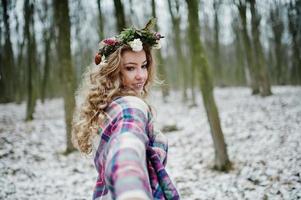 lockiges süßes blondes Mädchen mit Kranz im karierten Plaid am verschneiten Wald am Wintertag. foto