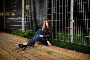 Nachtporträt von Mädchen Model Wear auf Jeans und Lederjacke gegen Eisenzaun. foto