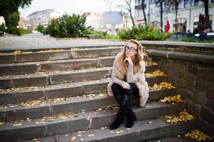 blondes mädchen im pelzmantel und brille, die auf treppen mit gelben blättern sitzen. foto