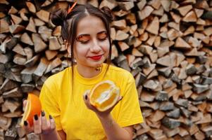 junges lustiges mädchen mit hellem make-up, tragen auf gelbem hemd halten stück orange gegen hölzernen hintergrund. foto