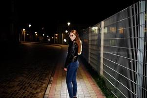 Nachtporträt von Mädchen Model Wear auf Jeans und Lederjacke gegen Eisenzaun. foto