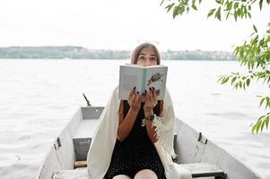 Porträt einer attraktiven Frau mit schwarzem Tupfenkleid, weißem Schal und Brille, die in einem Boot auf einem See ein Buch liest. foto