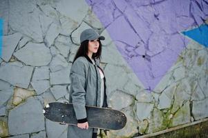 stilvolles brünettes mädchen in grauer kappe, lässiger streetstyle mit skateboard am wintertag gegen farbige wand. foto