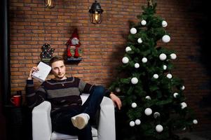 Studioporträt eines Mannes mit Buch, der auf einem Stuhl gegen einen Weihnachtsbaum mit Dekorationen sitzt. foto