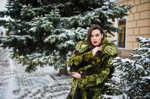 Brunettemädchen im grünen Pelzmantel am Wintertag gegen schneebedeckte Kiefer. foto