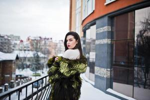 brünettes mädchen im grünen pelzmantel an der straße der stadt gegen haus mit großen fenstern im winter. foto