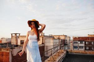 Porträt einer hübschen jungen Frau in weißem T-Shirt und blauem Rock, die mit ihrem orangefarbenen Hut bei Sonnenuntergang auf dem Dach posiert. foto