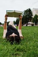 Porträt einer schönen Frau in schwarzem Tupfenkleid, die auf dem Gras liegt und ein Buch im Park liest. foto