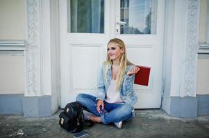 Blonde Mädchen tragen Jeans mit Rucksack, die mit Tagebuch gegen alte Tür gestellt wurden, und schreiben etwas. foto