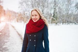 Blondes Mädchen in rotem Schal und Mantel, das am Wintertag im Park spazieren geht. foto