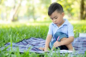 junger kleiner asiatischer junge liest und schreibt buch im park, asien kinderhausaufgaben und studium im sommer, kind entspannen sich beim zeichnen auf notizbuch im urlaub, bildungs- und entwicklungskonzept. foto