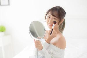schönheitsporträt junge asiatische frau lächelt mit gesicht, das spiegel beim schminken mit bürstenwange im schlafzimmer anwendet, schön von mädchen, die blusher, hautpflege und kosmetisches konzept halten. foto