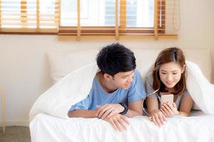 Junges asiatisches Paar lächelt, hört Musik mit Smartphone auf dem Bett im Schlafzimmer mit Spaß und genießt, Familie teilt Podcasts oder schaut Videos aus dem Internet online mit Entspannung, Lifestyle-Konzept. foto