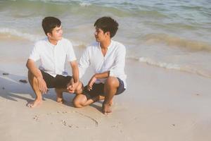 junges asiatisches paar schwul lächelnd romantisch herzform zusammen auf sand im urlaub zeichnend, homosexuell glücklich und spaß mit der liebe sitzend auf sand am strand im reisesommer, lgbt-rechtskonzept. foto
