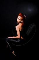 Mode-Modell rothaariges Mädchen mit ursprünglich Make-up wie Leoparden-Raubtier isoliert auf schwarz. Studioportrait auf Stuhl. foto