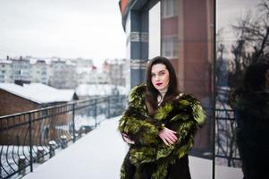 brünettes mädchen im grünen pelzmantel an der straße der stadt gegen haus mit großen fenstern im winter. foto