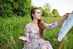 Porträt einer wunderschönen, glücklichen jungen Frau in schönem Kleid, die auf dem Gras sitzt und mit Wasserfarben auf Papier malt. foto