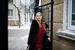 eleganz blondes mädchen in rotem abendkleid und pelzmantel auf den straßen der stadt im wintertag. foto