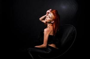 Mode-Modell rothaariges Mädchen mit ursprünglich Make-up wie Leoparden-Raubtier isoliert auf schwarz. Studioportrait auf Stuhl. foto