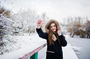 schönes brünettes Mädchen in warmer Winterkleidung. Modell auf Winterjacke gegen zugefrorenen See im Park, der Selfie am Telefon macht. foto