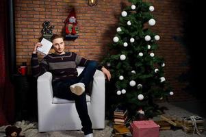 Studioporträt eines Mannes mit Buch, der auf einem Stuhl gegen einen Weihnachtsbaum mit Dekorationen sitzt. foto